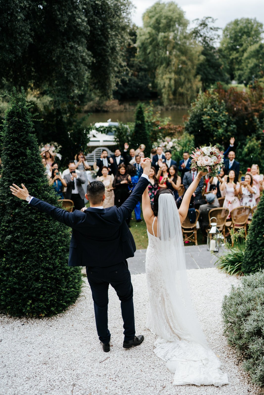 Bride and groom hold their hands up in celebration as guests look at them after outdoor ceremony at Bingham Riverhouse in Richmond