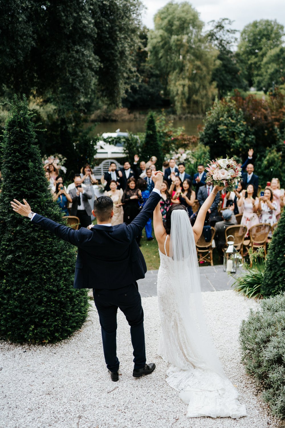 Bride and groom hold their hands up in celebration as guests look at them after outdoor ceremony at Bingham Riverhouse in Richmond