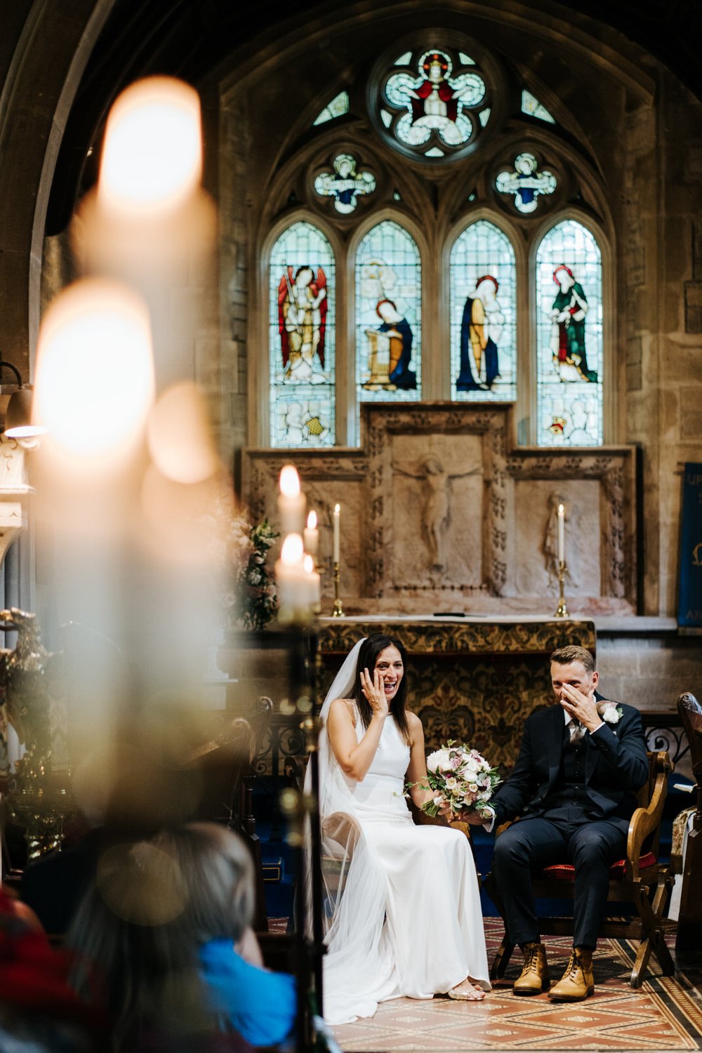 Bride and groom sit down and wipe tears off their eyes during English Cotswold church wedding in Lower Slaughter 