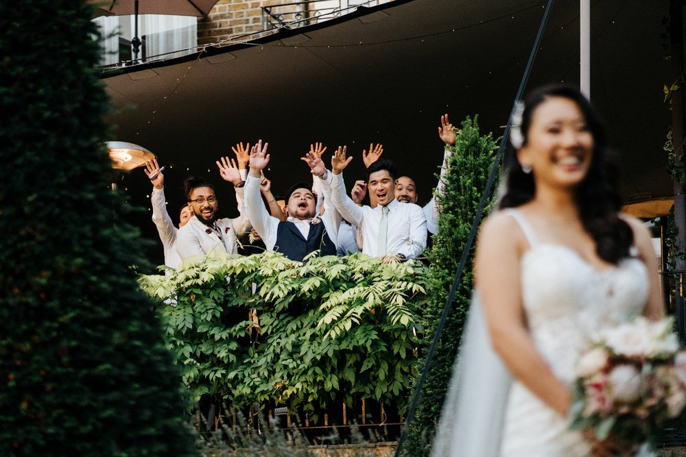 Bride, front and out of focus, smiles in embarrassment as groom's friends creep up from behind and cheer her on during photo session at Bingham Riverhouse