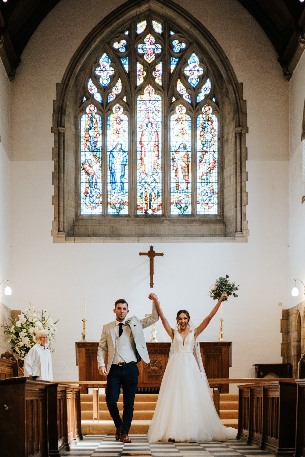 Bride and groom walk back down the aisle at church wedding while bride lifts her hands and bouquet in excitement 