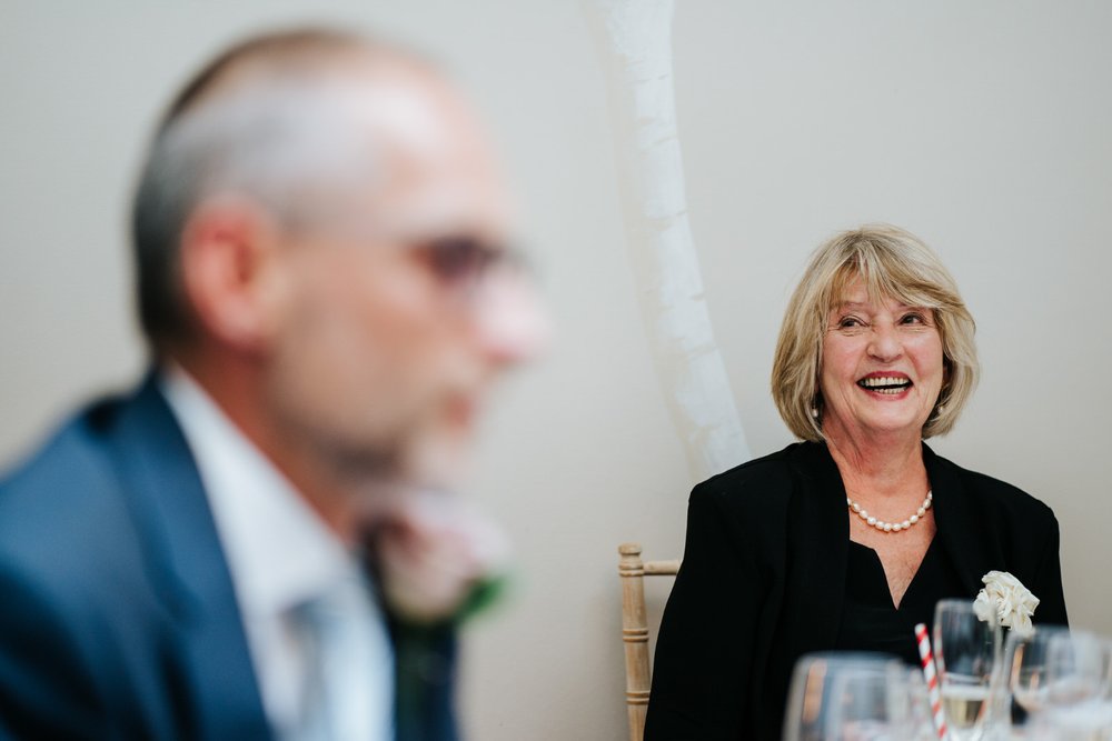 Mother of the bride smiles as her son-in-law, off frame, delivers wedding speech
