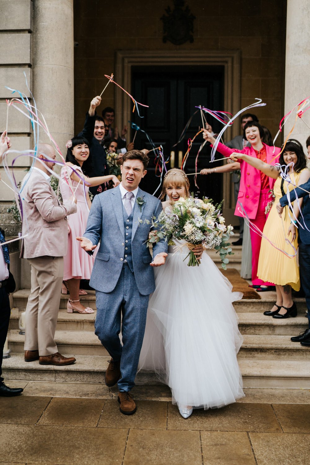 Bride and groom walk out of Hedsor House as guests wave colourful streamers