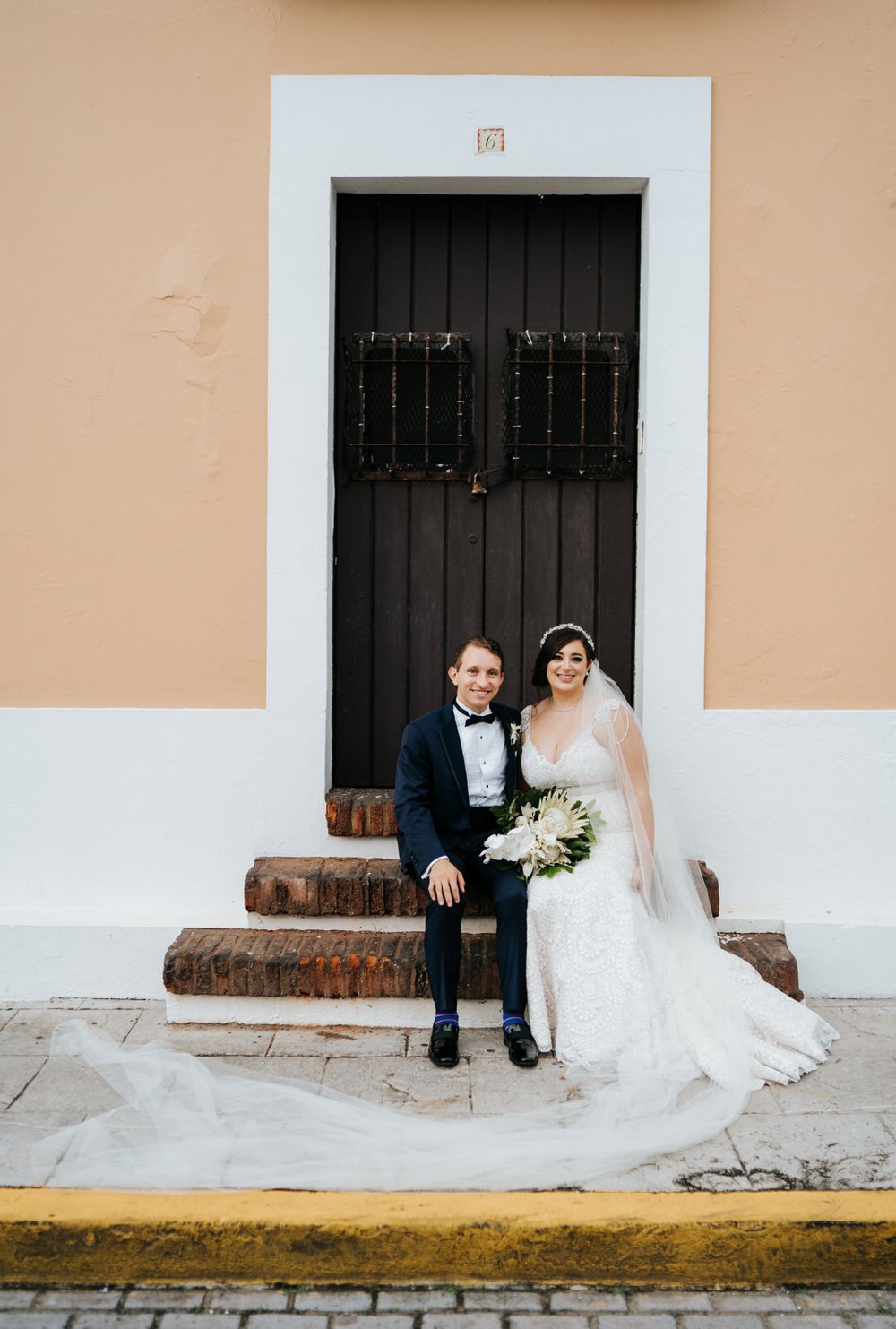 Bride and groom sit on steps of house in Old San Juan as bride's veil trails to the left during portraits taken on their wedding day 