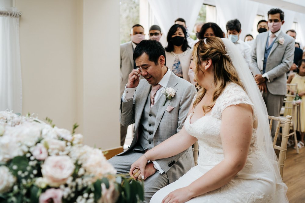 Bride and groom sit at registrar's table at Pembroke Lodge in Richmond as groom wipes tear of joy off his face 