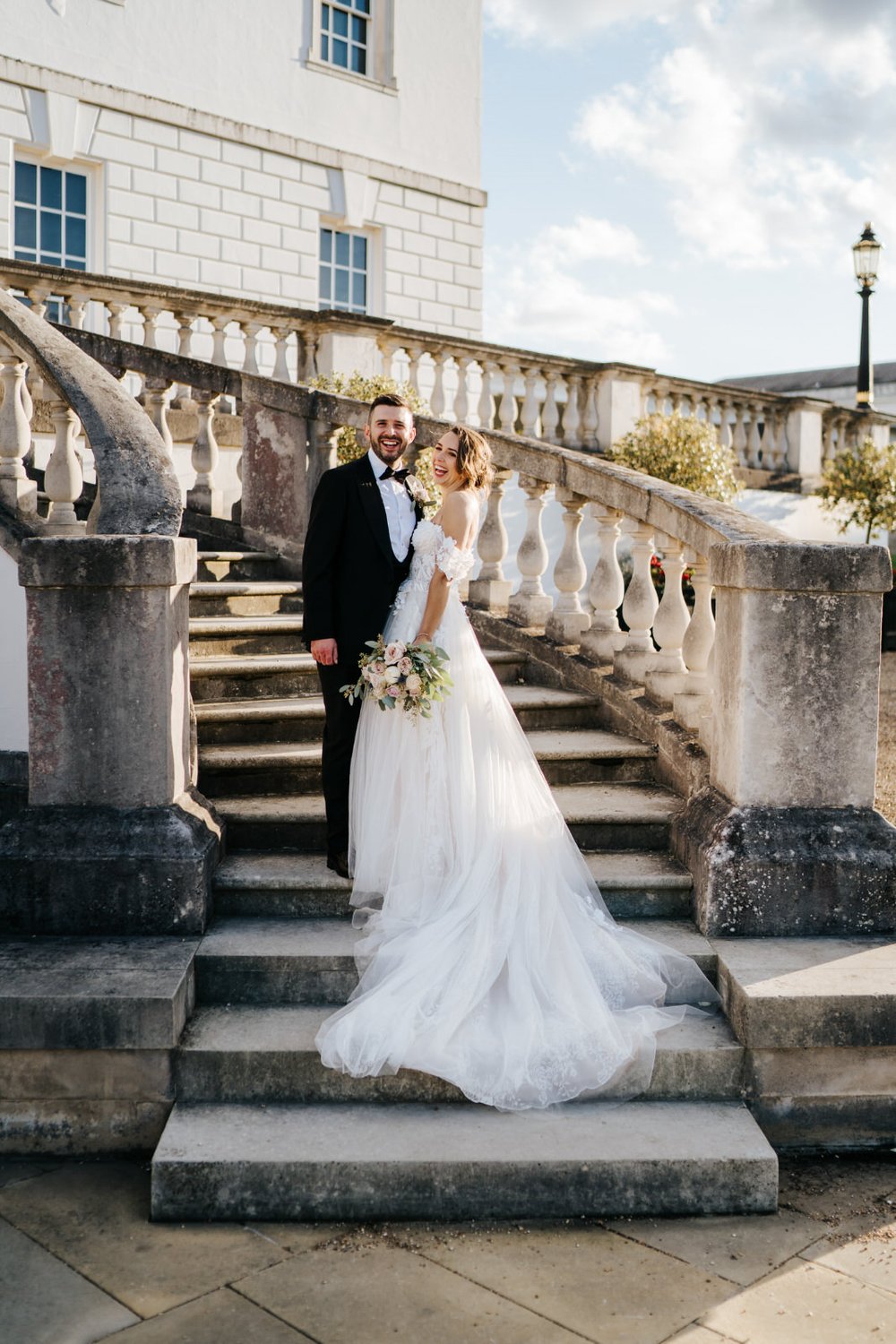 Bride and groom pose on stairs of The Queen's House wedding venue as sun sets off camera and bride's dress trails down stairs 