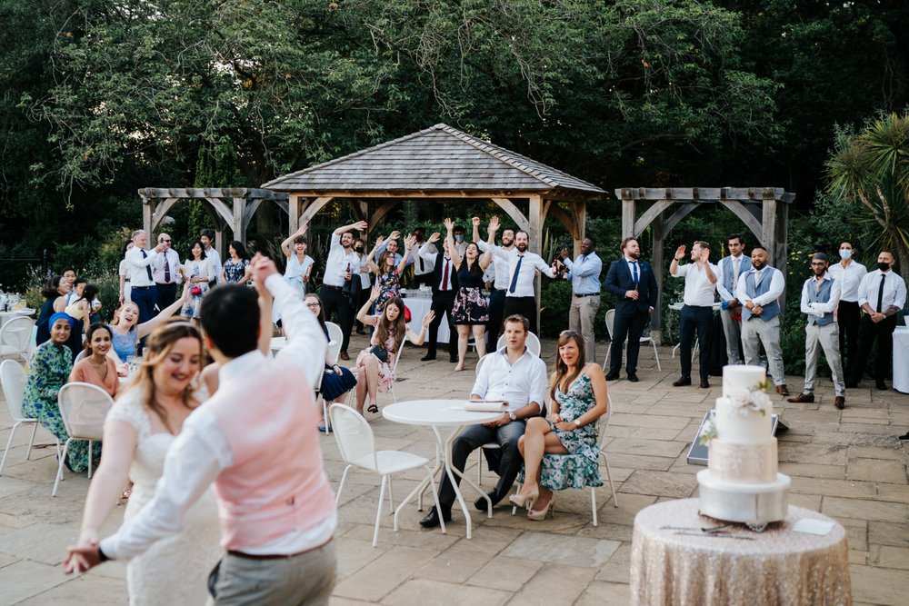 Wide photograph of outdoor first dance at Pembroke Lodge London as guests, in the background, wave their hands in the air