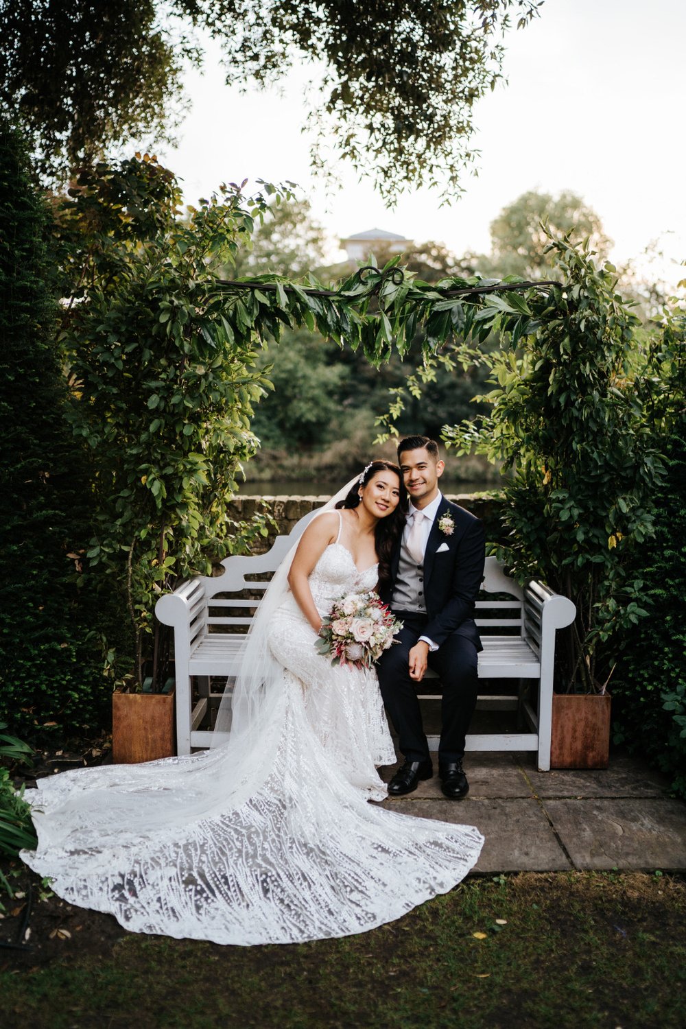 Bride and groom pose on bench outside Bingham Riverhouse while bride holds bouquet of King Protea by Johanna Pedrick Flowers