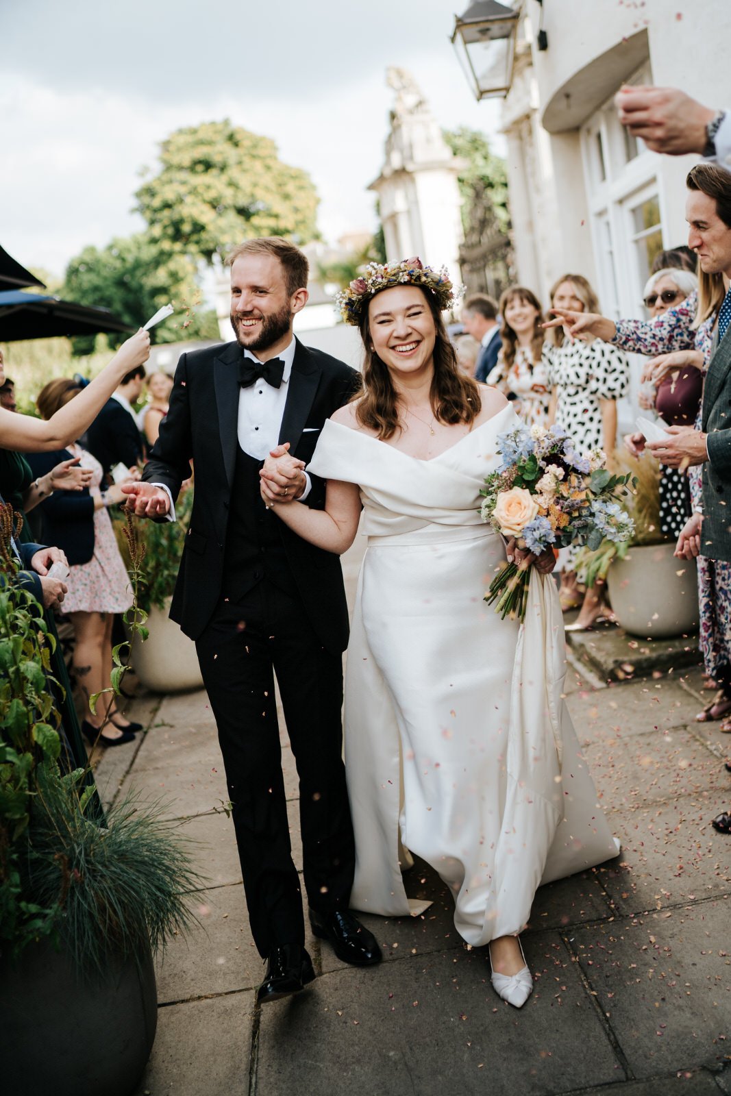 Bride and groom walk through tunnel of confetti at Hampton Court Palace wedding while bride holds beautiful flower bouquet by My Table Floral Design