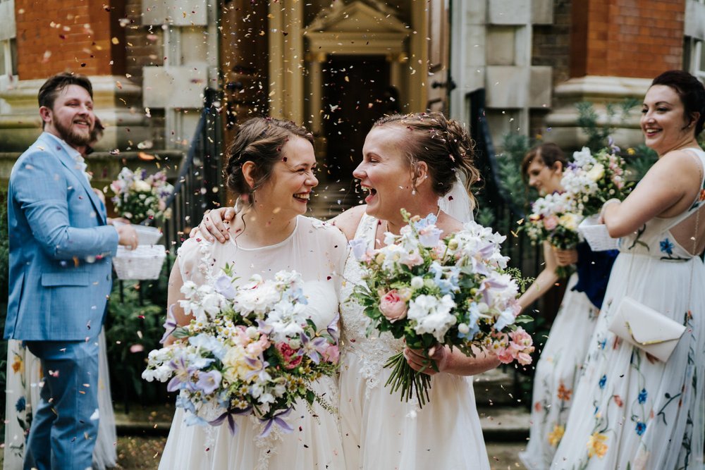 Two brides have confetti thrown at them as they walk out of Orleans House Gallery holding beautiful, colourful bouquets by Jessica Eliza Flowers