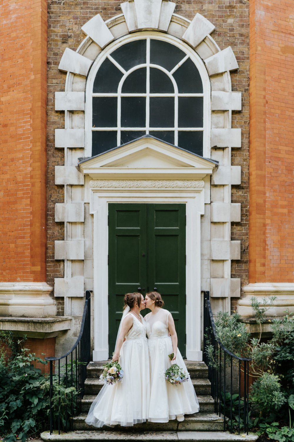 Two brides wearing white wedding dresses pose outside the doors of Orleans House Gallery