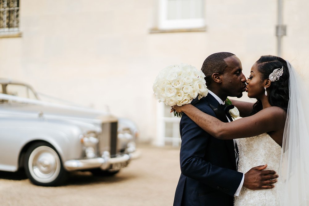 Bride and groom embrace and kiss while bride rests her round, white, rose bouquet on groom's shoulders with Rolls Royce in the background