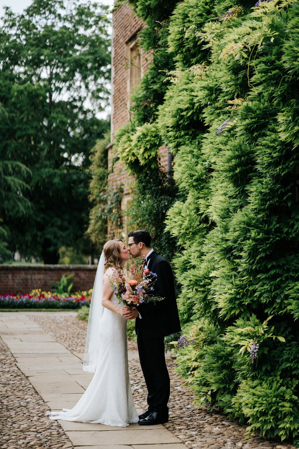Bride and groom pose in front of foliage at Jesus College in Cambridge while bride holds her colourful bouquet by Wild Rosamund