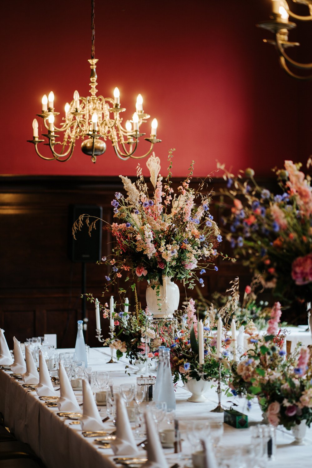Vase full of colourful and rustic flowers on top of head table at Jesus College in Cambridge with chandelier in background