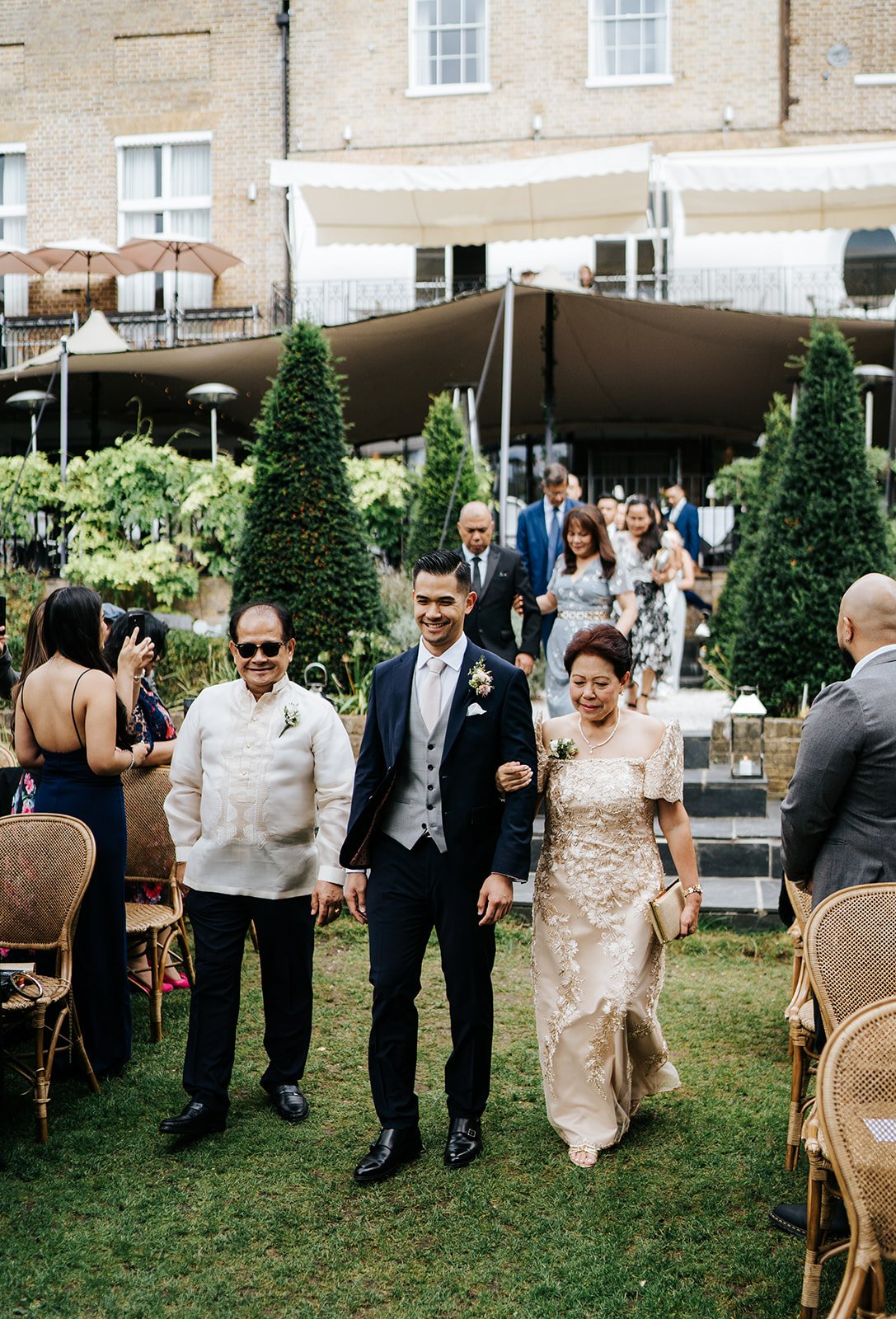 Groom walks down the aisle during outdoor ceremony at Bingham Riverhouse and is flanked by his mother and father