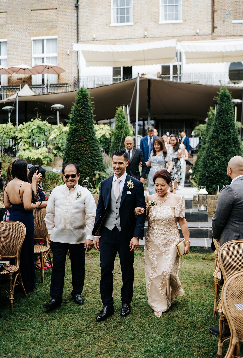 Groom walks down the aisle during outdoor ceremony at Bingham Riverhouse and is flanked by his mother and father