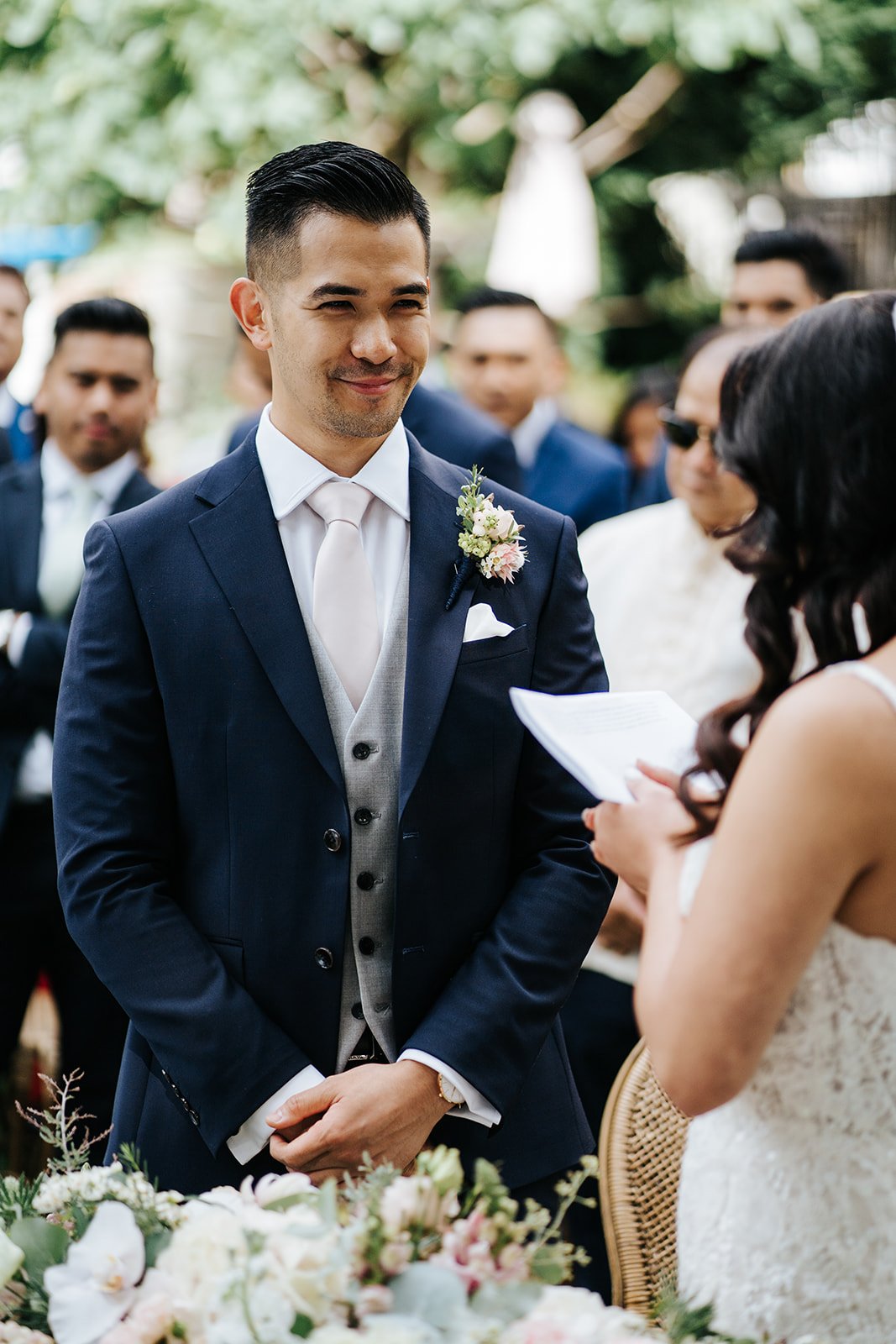 Groom stands and smiles in close-up photograph as bride reads her wedding vows to him