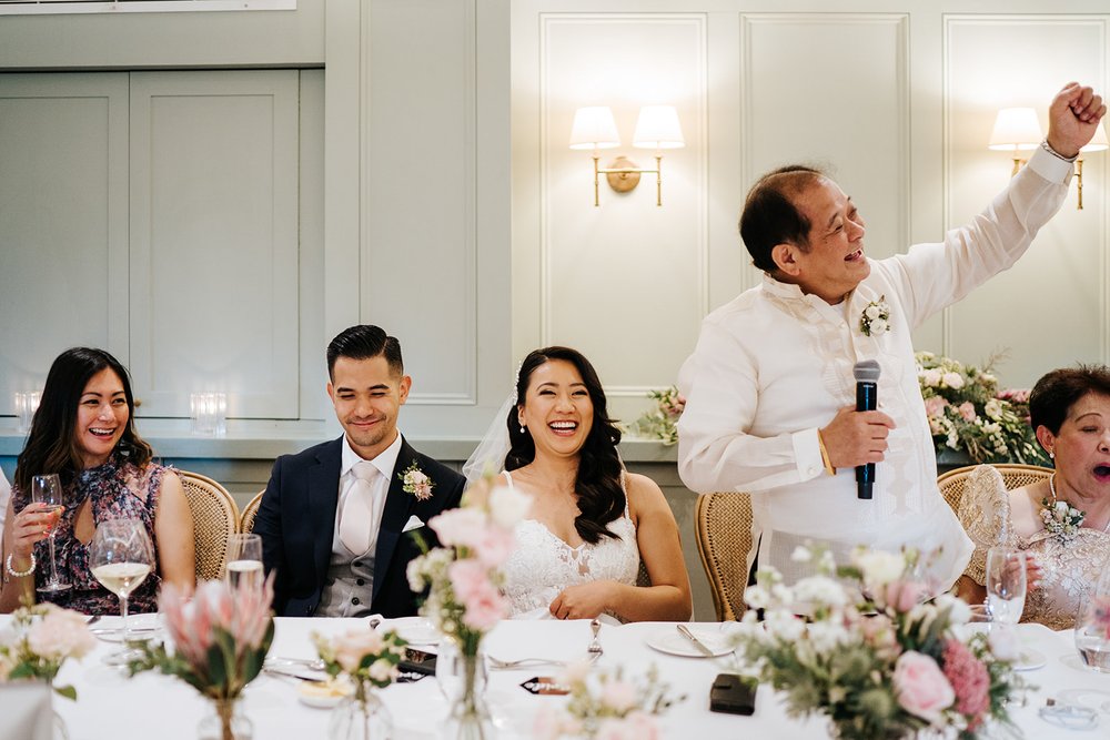 Father of the bride, to the right, delivers wedding speech and pumps fist in the air while bride and groom sit next to him and smile