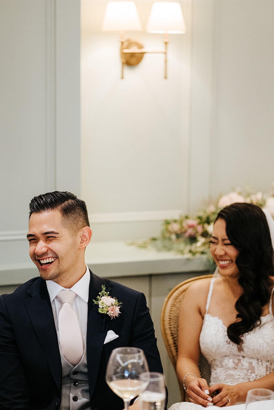 Close-up photo of bride and groom sat down during speeches and cackling with laughter
