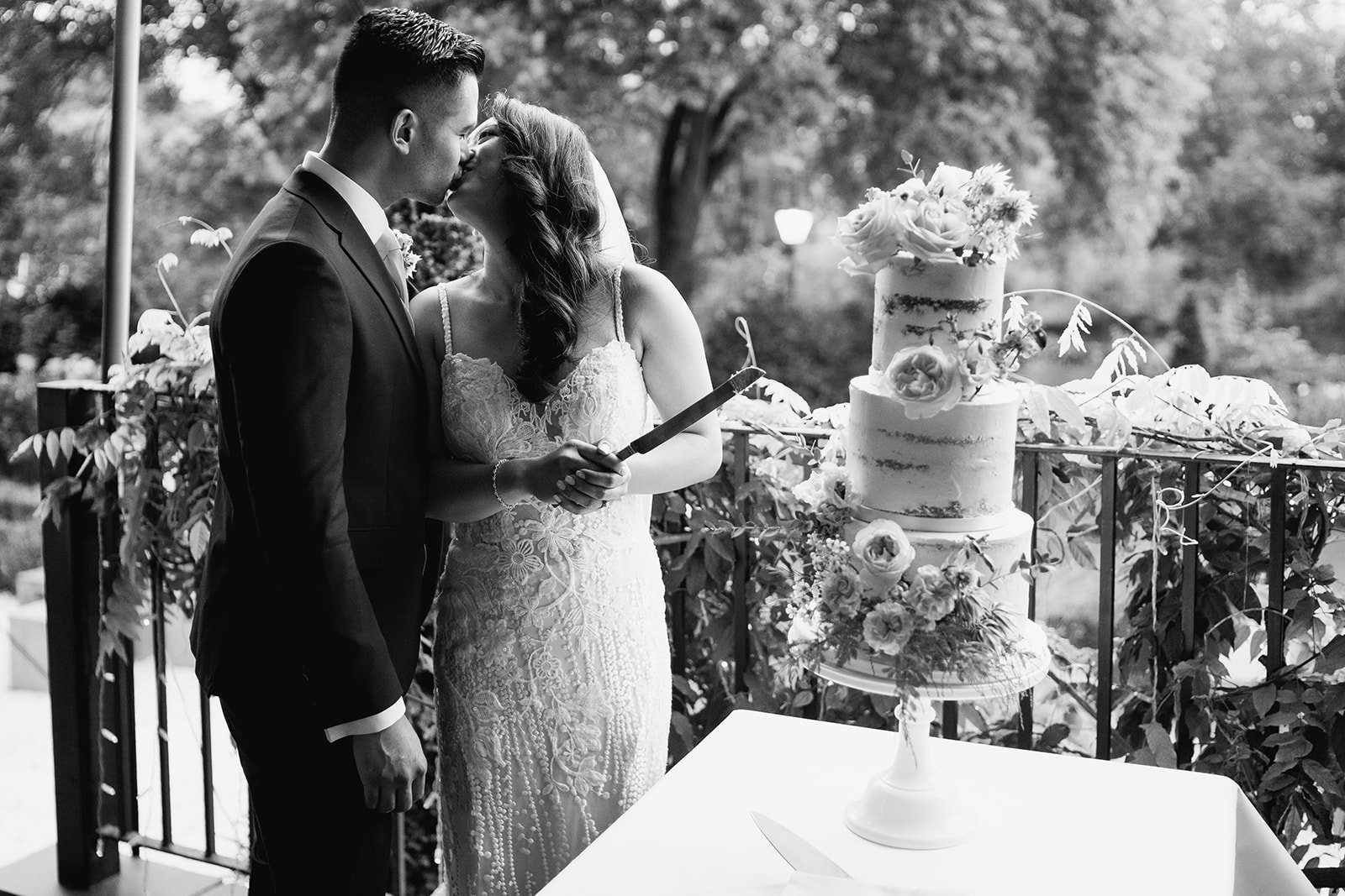Black and white photograph of bride and groom kissing after cutting beautiful wedding cake resting on table