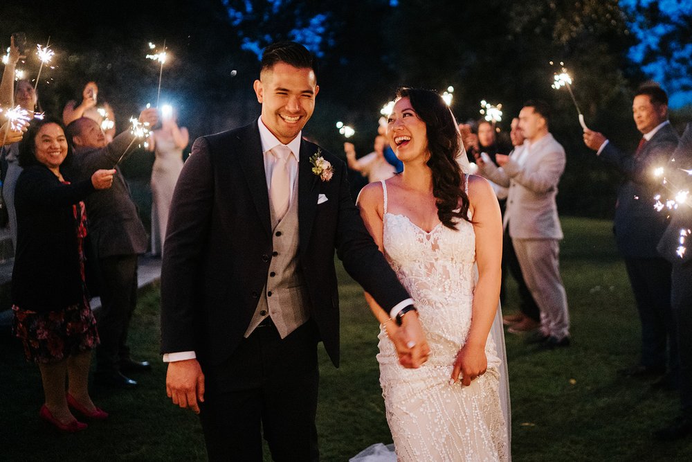 Bride and groom hold hands, look at each other and smile while being lit by guests holding sparklers next to them