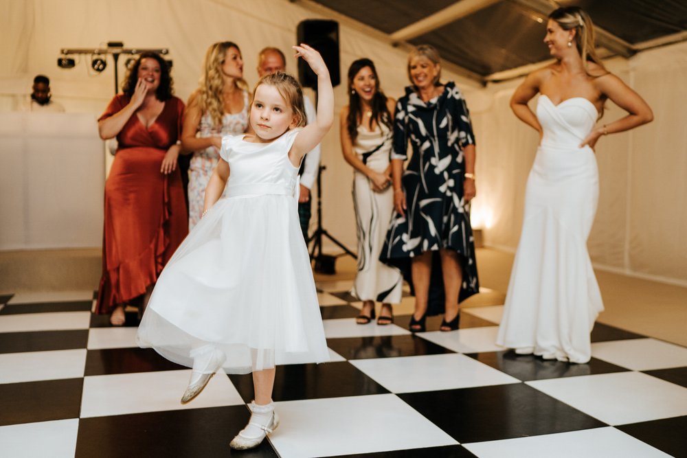 Little bridesmaid twirls on dancefloor as bride and her friends watch and cheer from behind at marquee wedding in Little Houghton