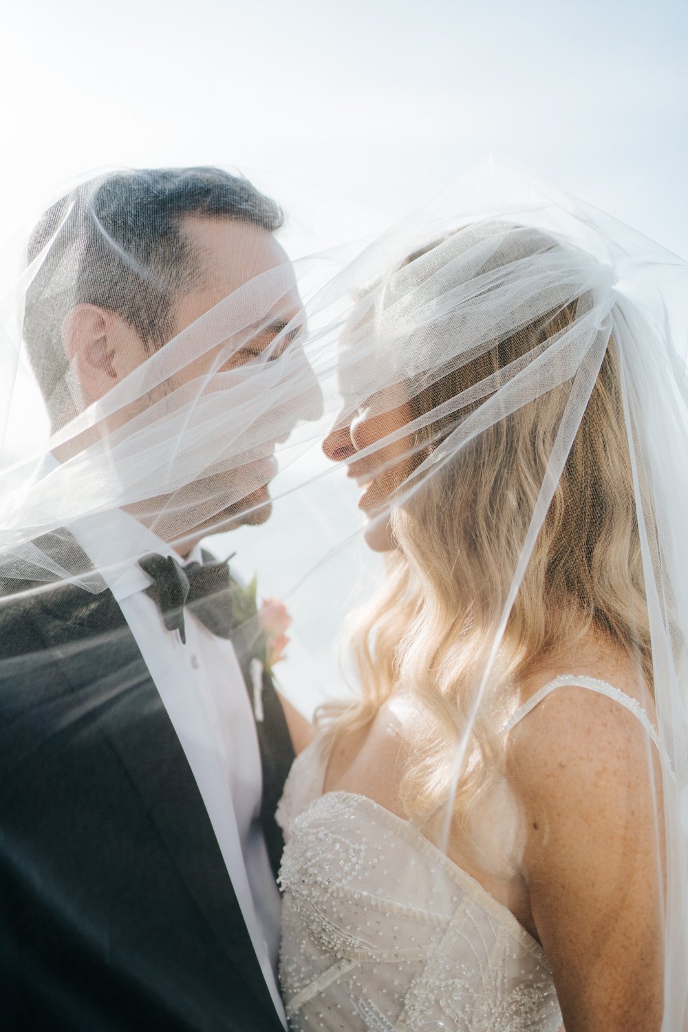 Bride and groom, draped in veil, smile in romantic capture at Puerto Rico beachfront wedding