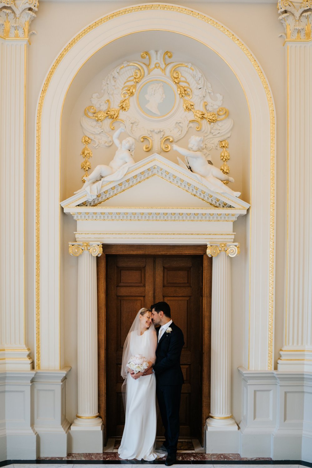 Bride and groom embrace lovingly in beautiful, ornate door frame of Orleans House Gallery