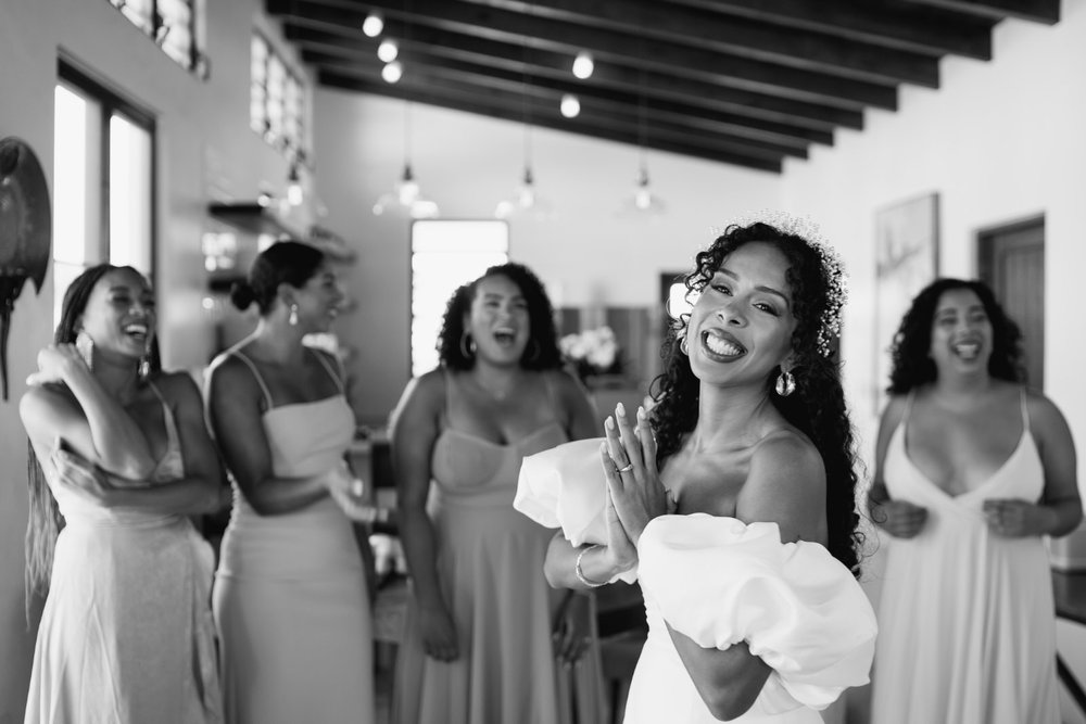 Bride, clasping hands and looking at camera with a smile as her bridesmaids react to seeing her in her wedding dress for the first time in Puerto Rico wedding in Vieques