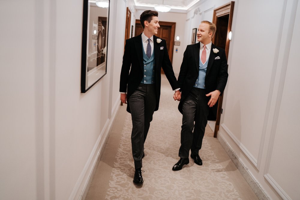Grooms, holding hands, walk down corridor of Claridge's Hotel as they direct themselves to wedding ceremony