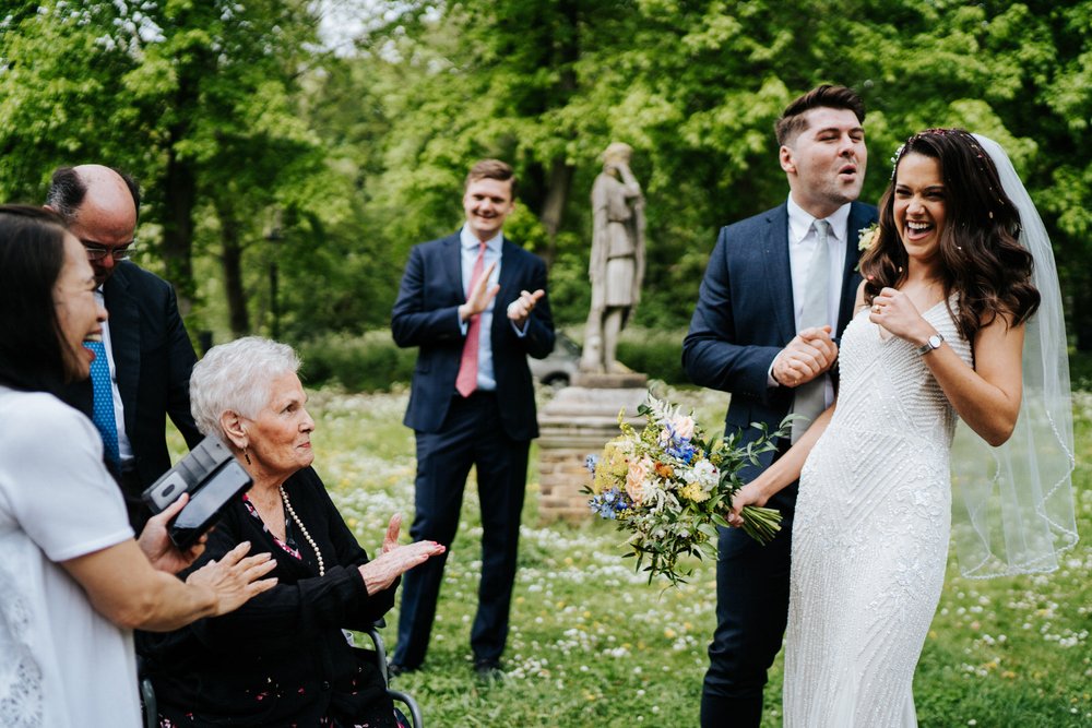 Bride reacts to her grandma pelting her with confetti while groom blows bride's hair to remove the confetti after wedding ceremony at Orleans House Gallery