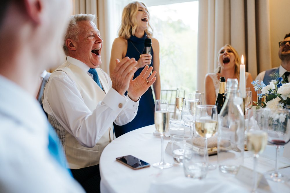 Father of the bride, sister of the bride and bride react with pure laughter to wedding speech at Pynes House wedding in Exeter