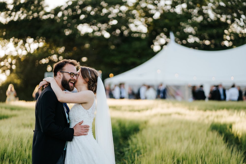 Groom smiles at camera as bride whispers something in his ear and sun sets in the back at Falmouth marquee wedding