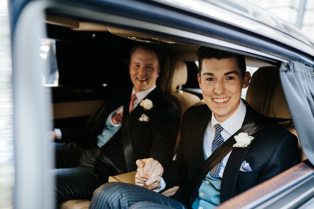 Two grooms sit in back of car, squeezing hands tightly, as they depart for Claridge's wedding reception in London