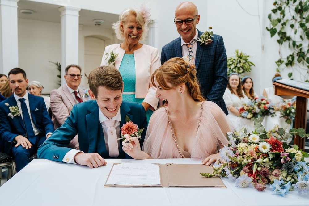 Bride and groom laugh as they sign their marriage certificate while their witnesses stand behind them at York House wedding
