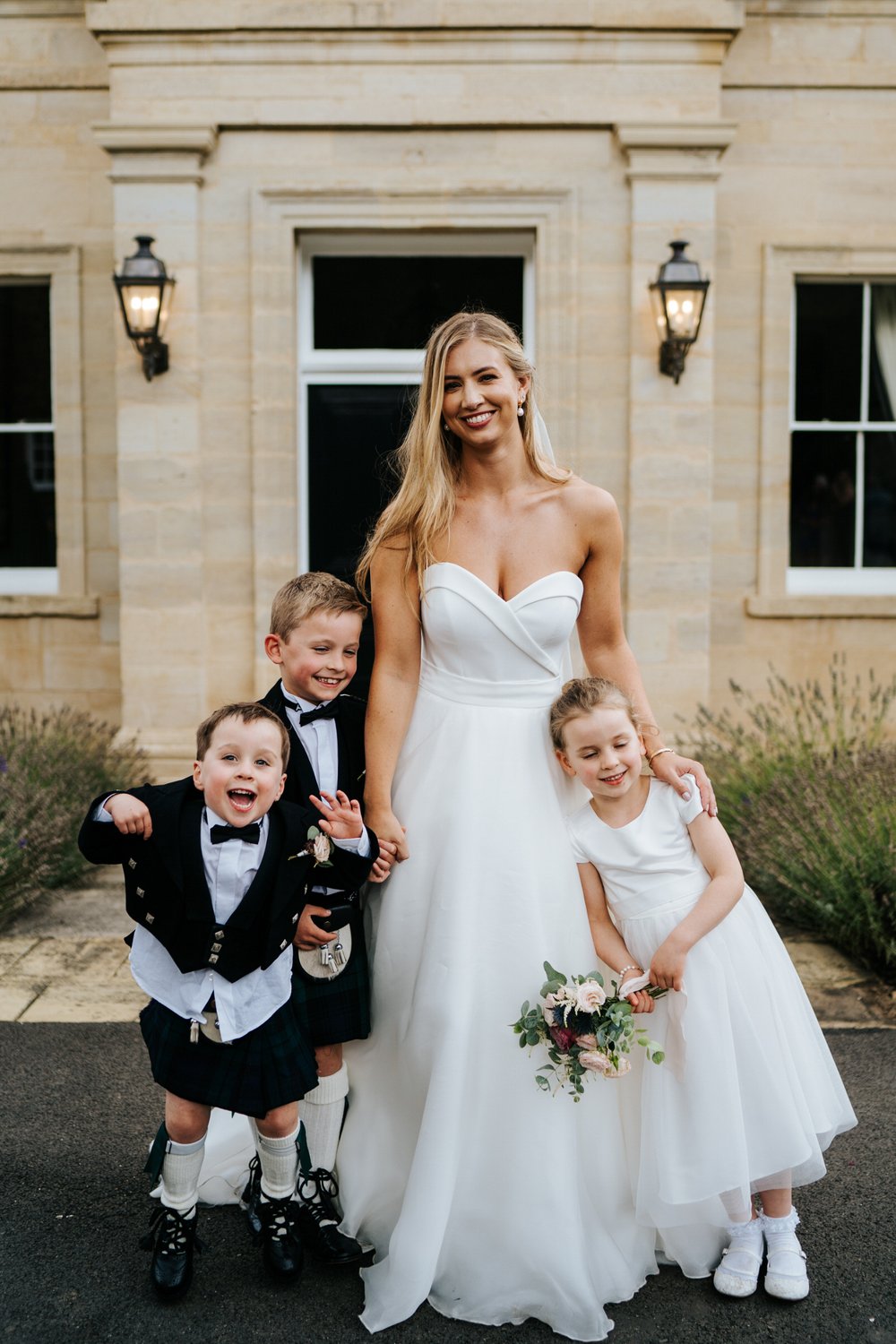 Bride poses with her three paigeboys and bridesmaids in front of Little Houghton house wedding