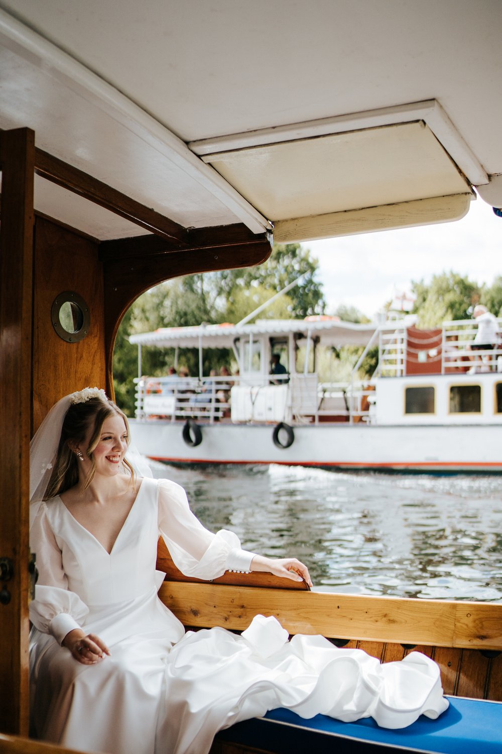 Bride sits on boat and smiles as she is taken to her wedding ceremony at Hampton Court House