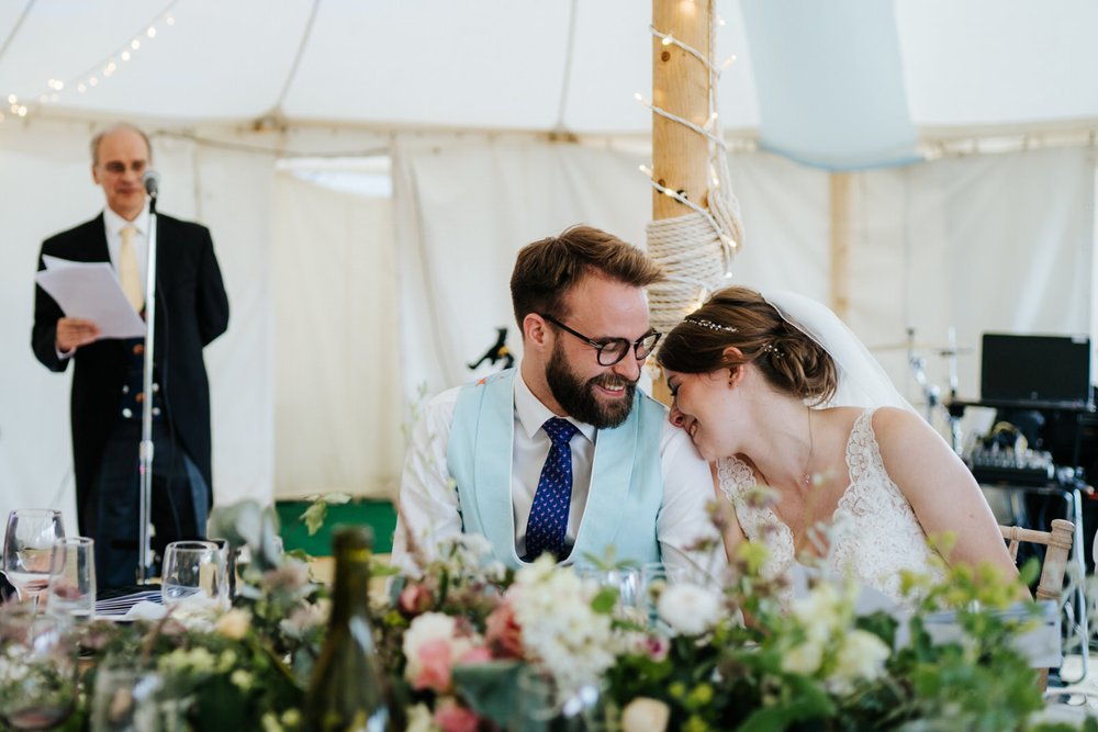 Bride leans her head on groom's shoulder in tender moment during wedding speeches at Falmouth marquee wedding