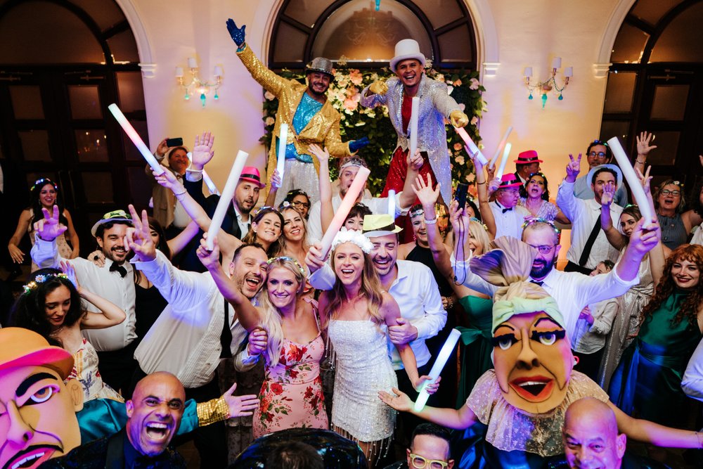 Wide group photograph of Puerto Rico wedding party as guests hold neon light batons and traditional Puerto Rican dancers wearing masks fill the frame