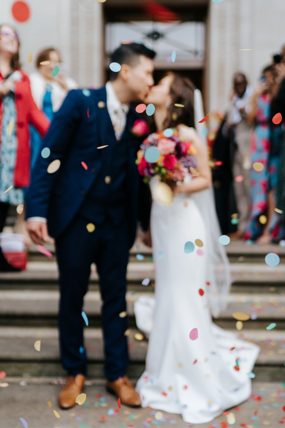 Bride and groom, out of focus, kiss while guests throw colourful, round confetti at them outside of Marylebone Town Hall in London