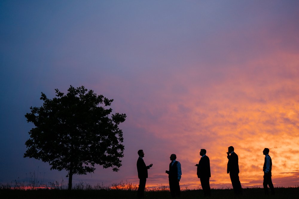 Beautiful sunset photograph with silhouetted groom and groomsmen enjoying a cigar at Chenies Manor House