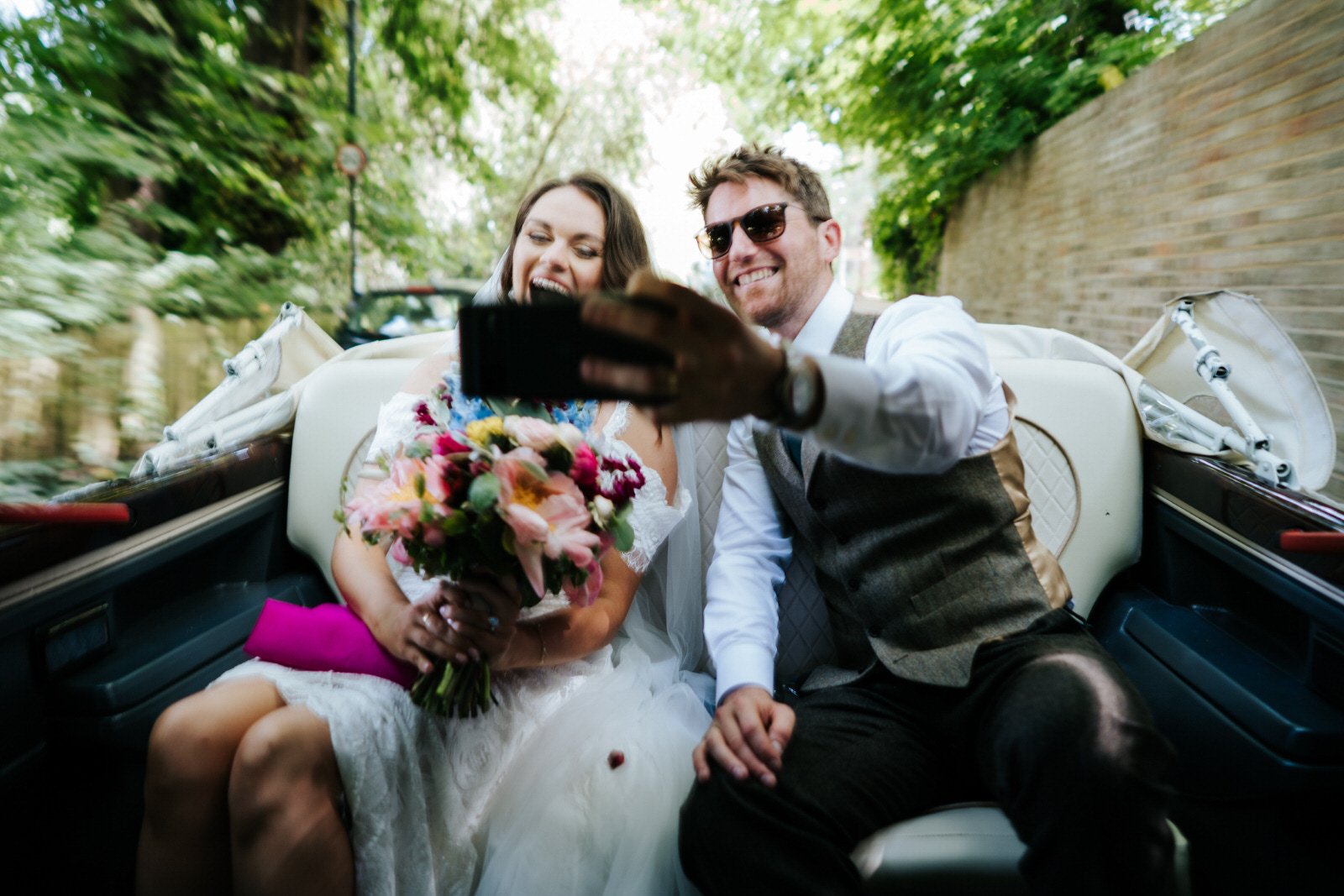 Blurry image of bride and groom taking a selfie as they depart their Orleans House Gallery wedding ceremony in car with no roof which will take them on joy ride across Richmond Park