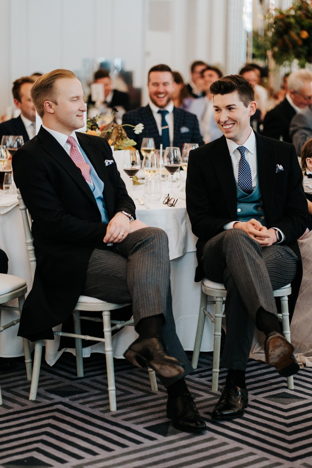 William Hanson and Michael Worrall sit cross-legged and smile at each other during wedding speeches at Claridge's