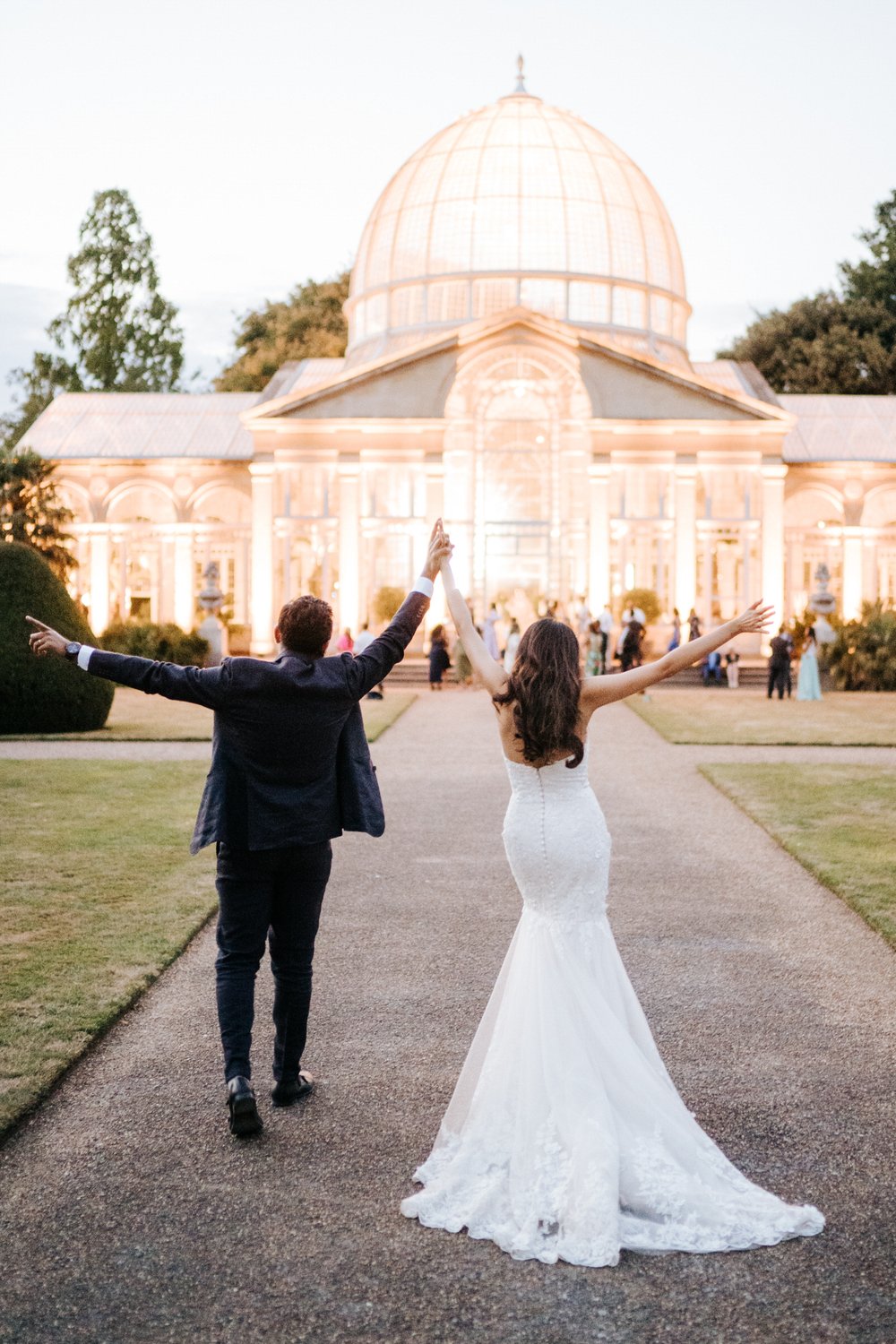 Bride and groom celebrate and throw their hands in the air as they walk back towards Syon House's Great Conservatory after spending some time taking photos