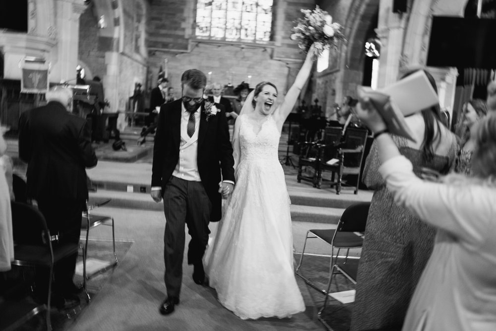 Bride and groom walk back down the aisle after church wedding as bride cannot control her excitement and waves her bouquet in the air