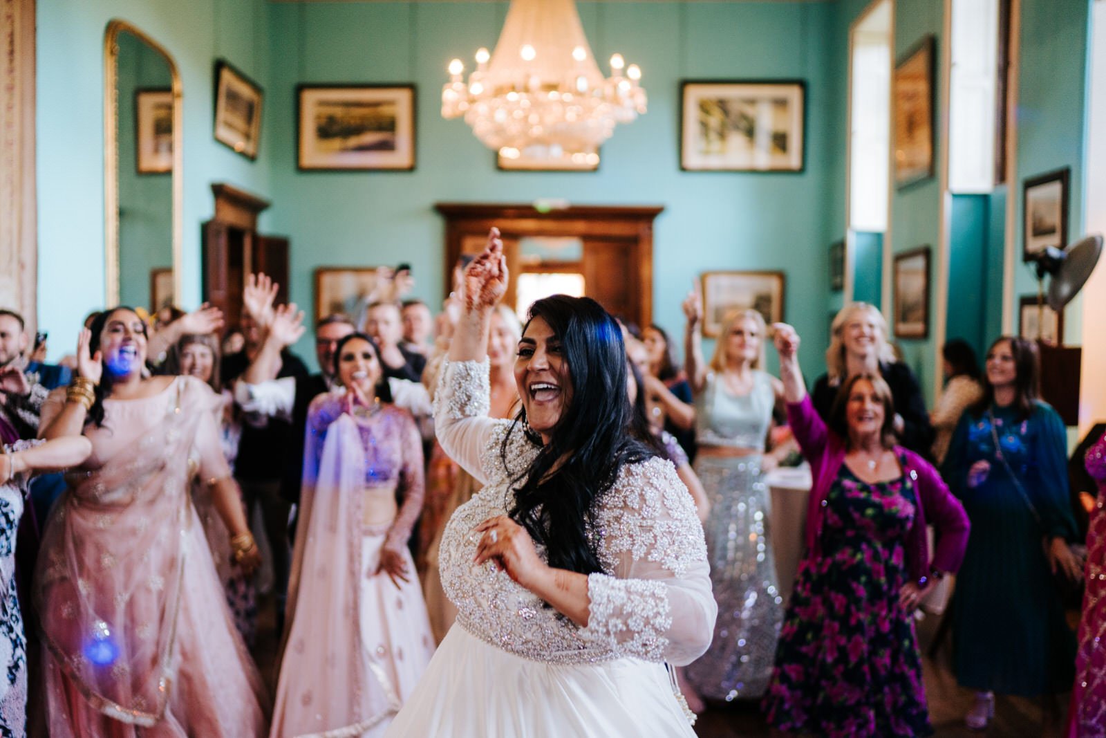 Bride, in middle of frame, throws one hand in the air as she dances Bollywood dance in Walcot Hall wedding