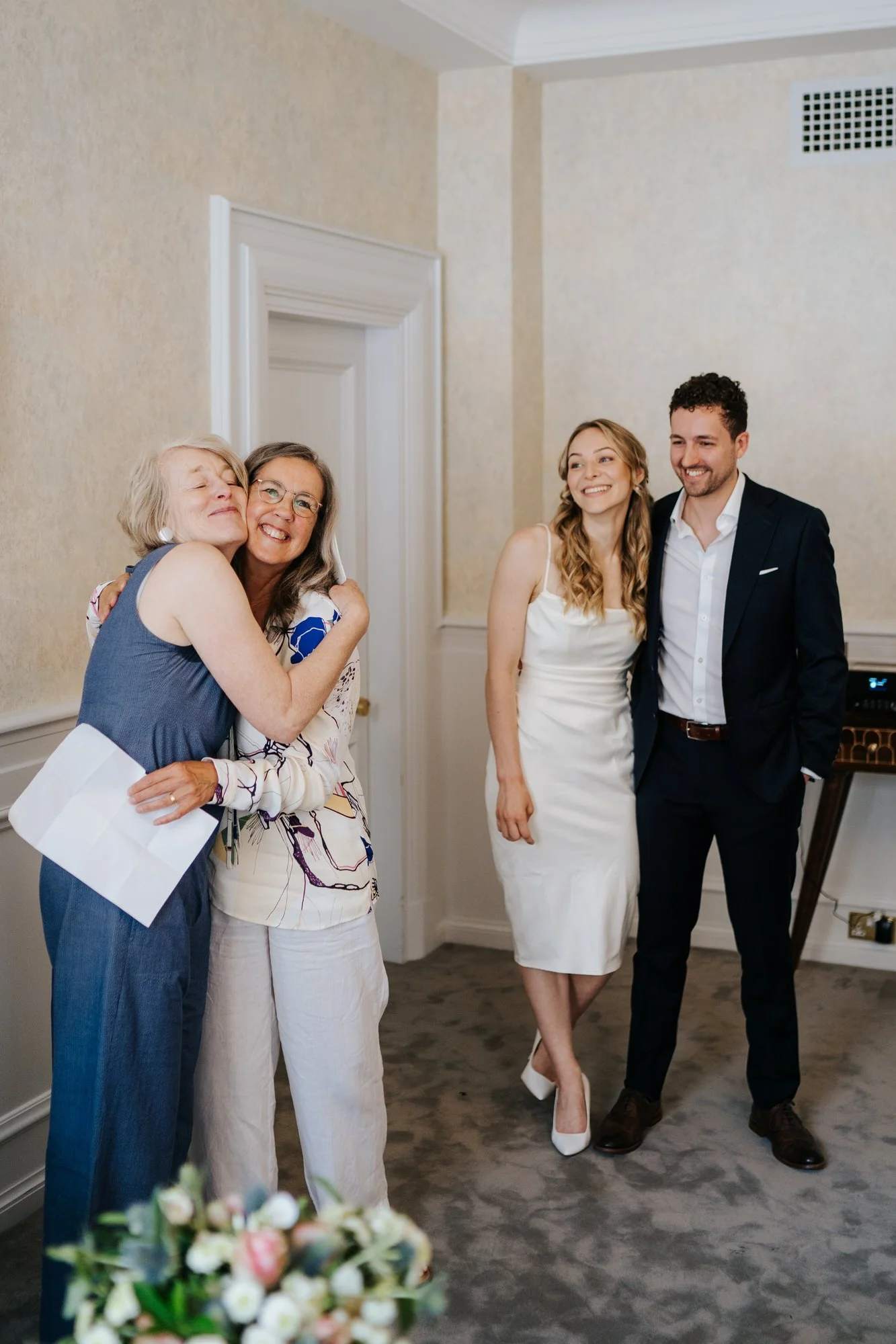 Mother of the bride and mother of the groom hug each other after finishing a reading as groom and bride stand in the back