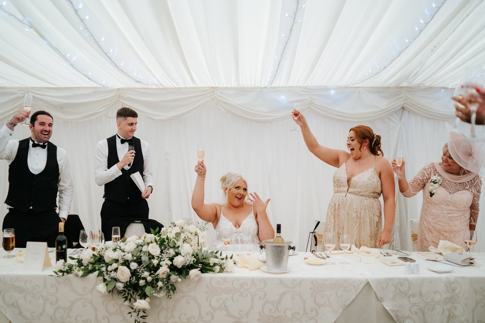 Bride, seated, raises her hand and shows of her wedding ring as all the guests around her clap and smile 