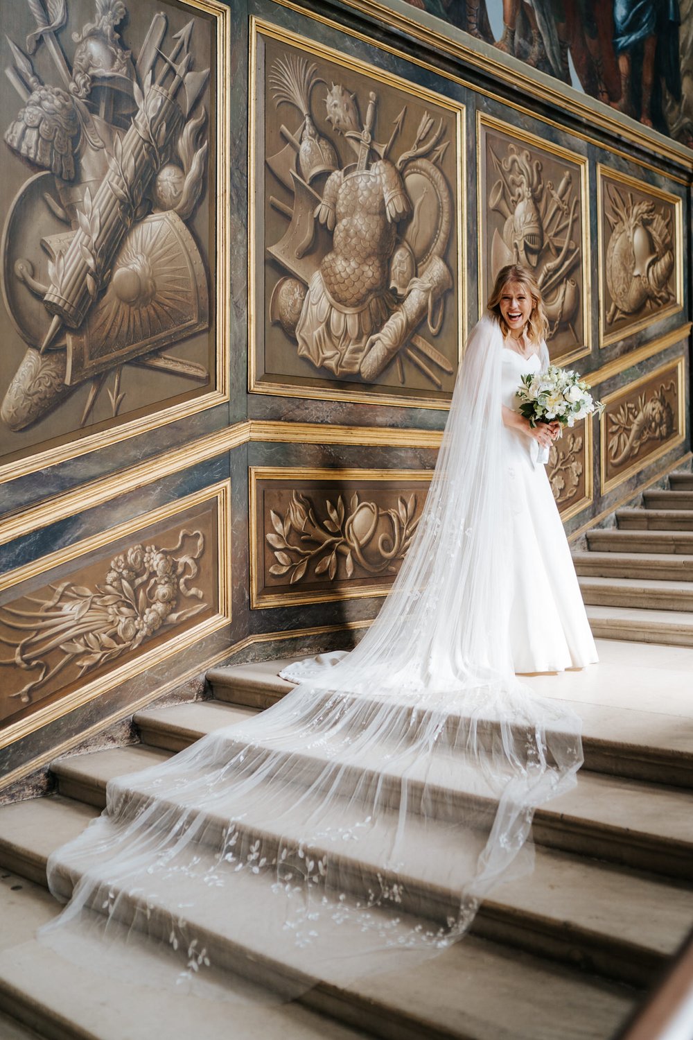 Bride cannot contain her joy and excitement as she stands in grand staircase at Hampton Court Palace while her veil flows behind her