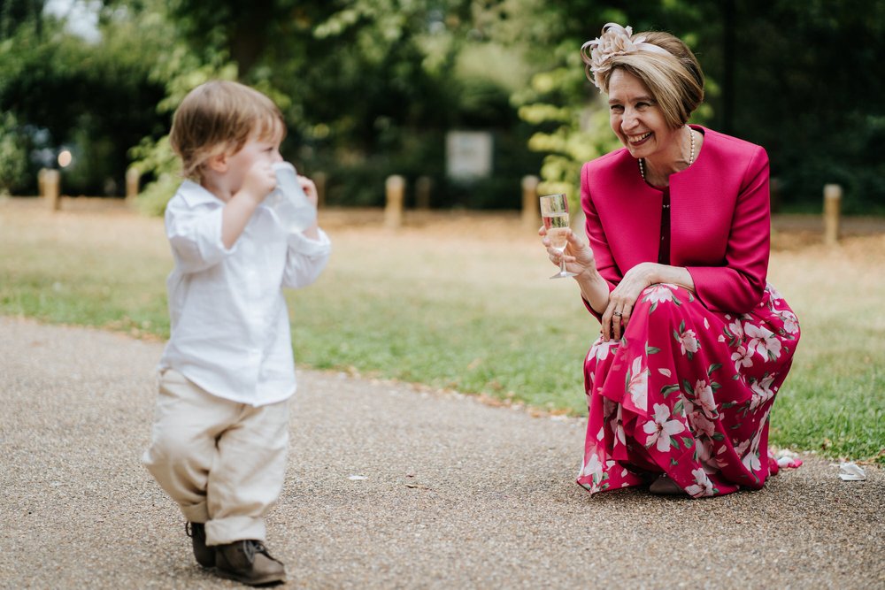 Mother of the bride, in elegant magenta dress, kneels with her champagne glass as little boy drinks from his water cup